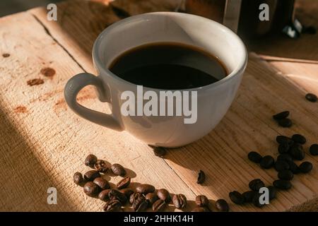 Fresh coffee in white ceramic cup near pile of freshly roasted coffee beans served on wooden table near French press Stock Photo