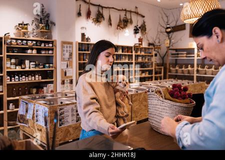 Young female with brown hair in casual clothe standing with small Chihuahua dog in hand in front of Woman seller and paying with smartphone in eco fri Stock Photo