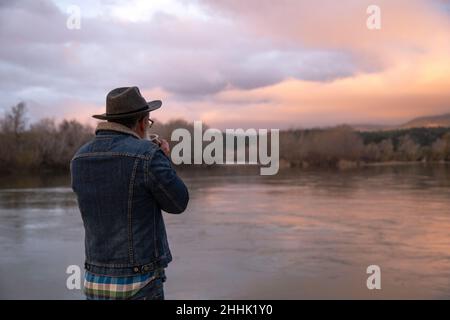 Back view of unrecognizable bearded male hiker in warm clothes drinking hot beverage while standing near lake during trekking on the sunset Stock Photo