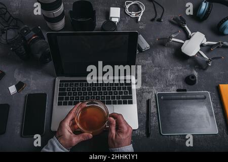 High angle of crop male photographer with netbook keyboard and drinking tea at table with graphic table photo camera smartphone batteries and drone ne Stock Photo