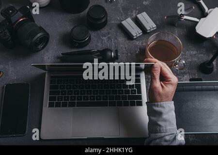 High angle of crop male photographer with netbook keyboard and drinking tea at table with graphic table photo camera smartphone batteries and drone ne Stock Photo