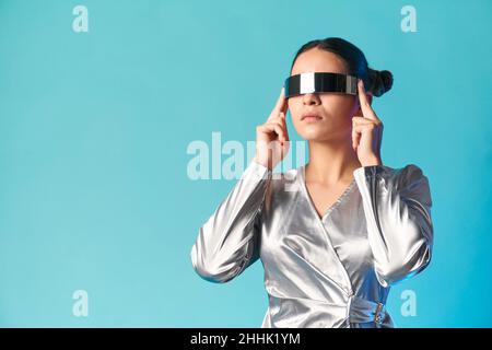 Concentrated young ethnic female model with dark hair in stylish dress adjusting futuristic VR goggles against blue background Stock Photo