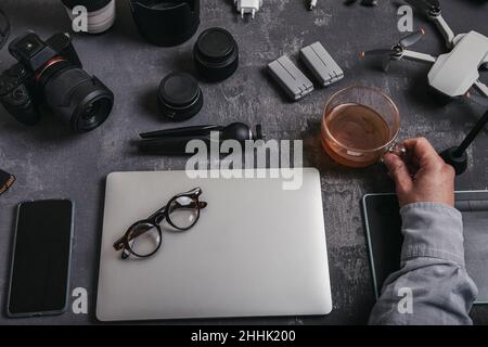 High angle of crop male photographer with netbook keyboard and drinking tea at table with graphic table photo camera smartphone batteries and drone ne Stock Photo