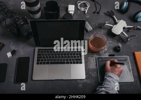 High angle of crop anonymous male photographer with stylus using graphic tablet at desk with laptop smartphone and devices for photo camera and UAV dr Stock Photo