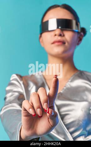 Low angle of stylish young ethnic lady with dark hair in trendy mirrored VR glasses touching invisible screen with finger while experiencing virtual r Stock Photo