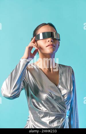 Concentrated young ethnic female model with dark hair in stylish dress adjusting futuristic VR goggles against blue background Stock Photo