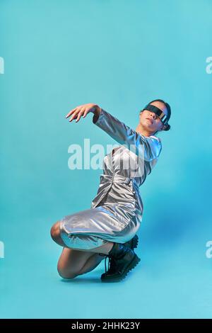 Side view of fashionable young ethnic female in futuristic outfit and VR goggles sitting on haunches and gesturing while exploring virtual reality in Stock Photo