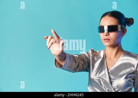 Side view of stylish young ethnic lady with dark hair in trendy mirrored VR glasses touching invisible screen with finger while experiencing virtual r Stock Photo