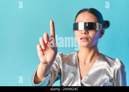 Stylish young ethnic lady with dark hair in trendy mirrored VR glasses touching invisible screen with finger while experiencing virtual reality agains Stock Photo