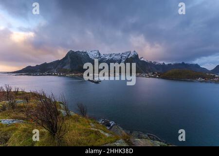 Reine village located on island surrounded with calm blue water against snowy mountains and cloudy sundown sky Stock Photo