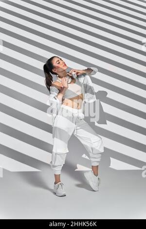 Full body of female dancer in sportswear performing freestyle dance with wireless headphones while enjoying music in studio with stripes of shadows du Stock Photo