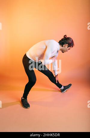 Full body of fit young Hispanic sportsman with dark hair in activewear doing stretching exercise for legs while standing against orange background dur Stock Photo