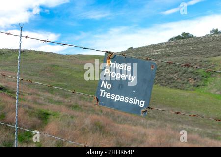 Private Property, No trespassing sign on barbed wire fence. Blurred green hills background under blue sky. Stock Photo