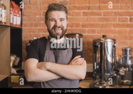 Bearded positive male barista in apron crossing arms while standing in coffee house and looking at camera Stock Photo