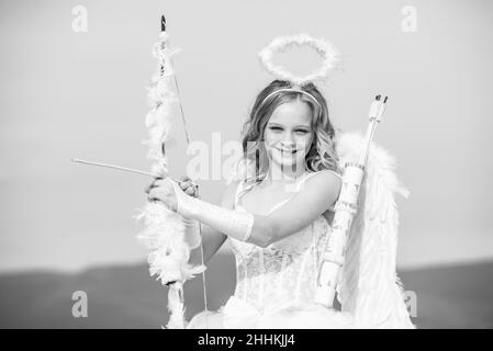 Innocent Girl with angel wings standing with bow and arrow against blue sky and white clouds. St Valentines day. Little angel girl against sunny sky Stock Photo