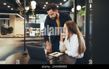 Focused businesspeople collaborating on a project. Two young entrepreneurs using a laptop while working together in a modern workspace. Two young busi Stock Photo