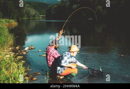 Male hobby fishing. Two male friends dressed in shirts fishing together with net and rod during the morning light on the lake. Man at riverside enjoy Stock Photo