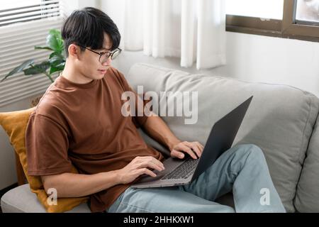 Technology Concept The man that is putting on glasses leaning his back on the dark yellow while focusing on the laptop’s screen. Stock Photo