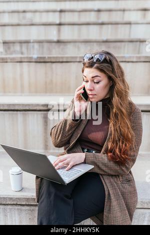 Female freelancer in outerwear with long hair. browsing data on netbook and speaking on cellphone while sitting on stairs near coffee to go on city street Stock Photo