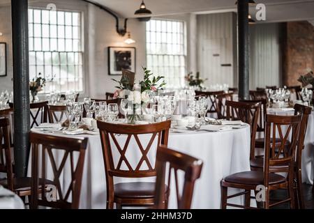 Romantic wedding breakfast room set up for guests and the bride and groom.  Wooden chairs, tables, glasses flowers and industrial setting. Stock Photo