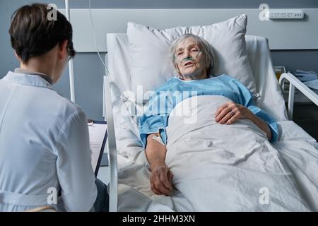 Senior woman lying on bed at the ward and talking about her disease with the nurse Stock Photo
