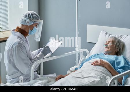 Young doctor in protective wear writing prescription in medical card while visiting senior woman at hospital ward Stock Photo