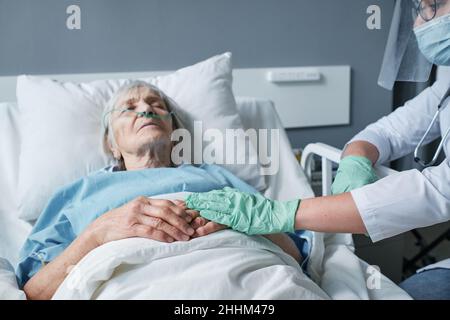 Doctor in protective gloves holding hands of senior woman while she lying on bed at ward, doctor caring about her during disease Stock Photo
