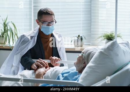 Young son in protective mask visiting his sick senior mother at hospital ward during pandemic Stock Photo