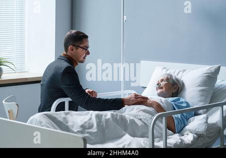 Young man talking to her sick mother during his visit at hospital, she lying on bed and smiling Stock Photo