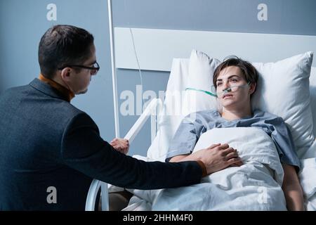 Rear view of young man supporting the sick young woman while she lying on bed at hospital ward Stock Photo