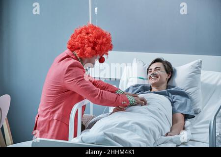 Happy young woman having fun with the clown while lying on hospital bed during her treatment Stock Photo