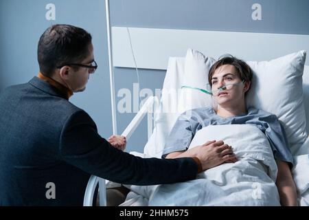 Rear view of young man supporting the sick young woman while she lying on bed at hospital ward Stock Photo