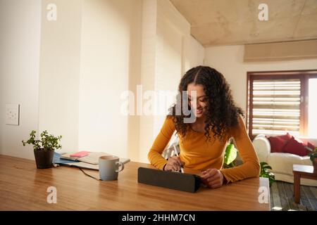Multi-cultural young adult female smiling while using using her electronic tablet at her modern kitchen counter Stock Photo