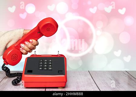 A feminine elegant beautifully manicured woman's hand holding an old red telephone handset over an abstract blurry light pink Valentine's Day or weddi Stock Photo