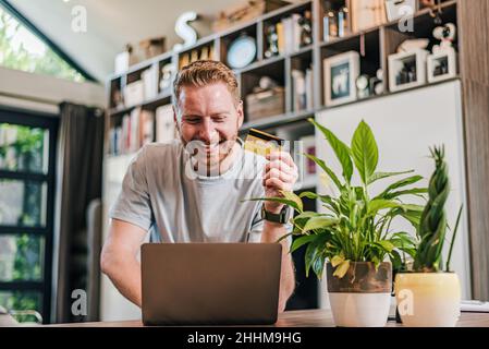 Happy cheerful smiling young adult man doing online shopping or e-shopping satisfied entrepreneur making online payment paying for service or goods ha Stock Photo