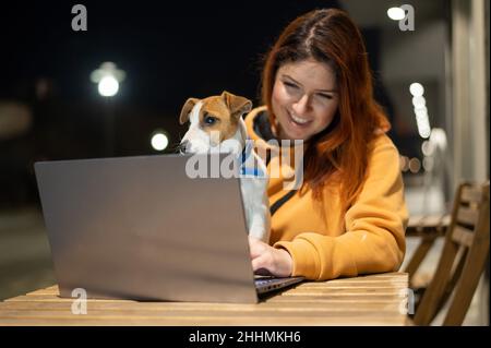 Smiling woman working on laptop at a wooden table in the street. The girl looks at the monitor with her pet jack russell terrier. Freelancer walks the Stock Photo