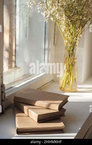 Stack of books and vase of white flowers stand on the windowsill by the window on sunny day. Stock Photo