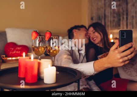 Valentines Day loving couple taking selfie on phone by wine glasses with strawberries, candles having romantic dinner celebrating at home at night Stock Photo