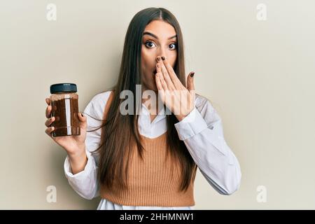 Beautiful brunette young woman holding soluble coffee covering mouth with hand, shocked and afraid for mistake. surprised expression Stock Photo