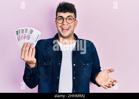 Young hispanic man holding egyptian pounds banknotes celebrating achievement with happy smile and winner expression with raised hand Stock Photo