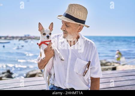 Senior man wearing summer hat hugging chihuahua at seaside Stock Photo