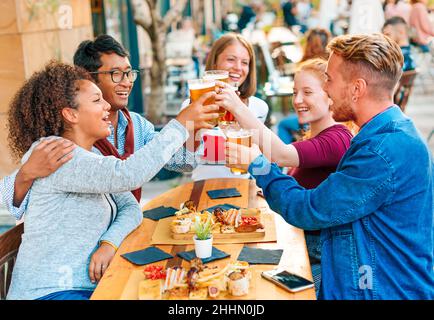 Satisfied multiracial friends sitting at table and looking at each other while clinking glasses with beer in street cafeteria Stock Photo