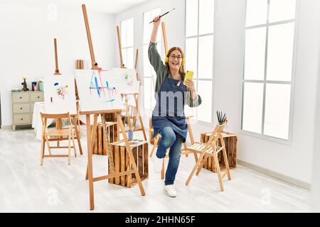 Middle age artist woman smiling happy and dancing painting at art studio. Stock Photo
