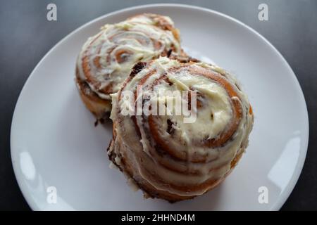 Close-up of two classic cinnamon rolls with cheese cream on top on a large white plate on a dark gray, almost black background. Stock Photo