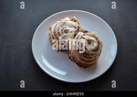 Close-up of two classic cinnamon rolls with cheese cream on top on a large white plate on a dark gray, almost black background. Stock Photo