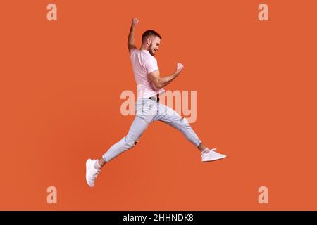 Side view portrait of happy bearded man jumping in air showing yes i did it gesture, copy space for ad, wearing pink T-shirt and jeans. Indoor studio shot isolated on orange background. Stock Photo