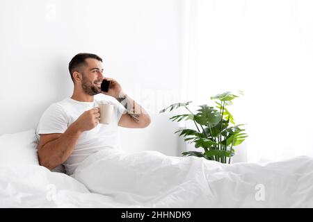 Happy young caucasian muscular man sit on white bed drink coffee and talk by phone in bedroom interior Stock Photo