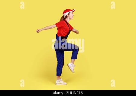 Side view portrait of worker woman marching, going for work, expressing positive emotions and happiness, wearing overalls and red cap. Indoor studio shot isolated on yellow background. Stock Photo