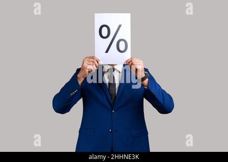 Portrait of anonymous unknown businessman hiding his face behind paper with percent sign inscription, wearing official style suit. Indoor studio shot isolated on gray background. Stock Photo