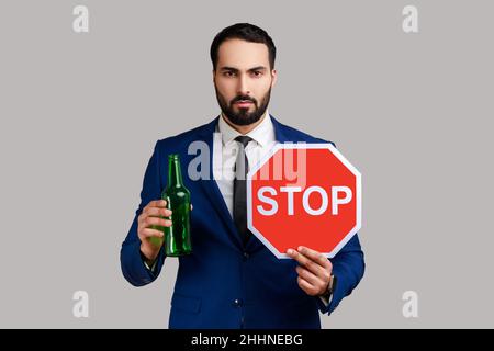 Portrait of serious strict bearded man showing alcoholic beverage beer bottle and stop sign, warning and worrying, wearing official style suit. Indoor studio shot isolated on gray background. Stock Photo